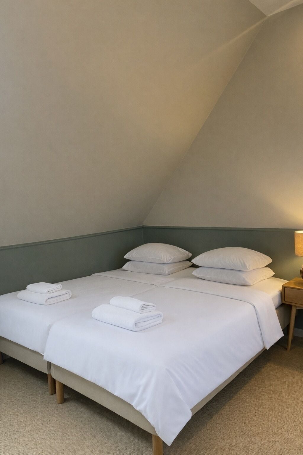 Una cama individual con sábanas y almohadas blancas, una mesita de noche de madera con una lámpara, y suelo cubierto de alfombra en una habitación con techo inclinado.