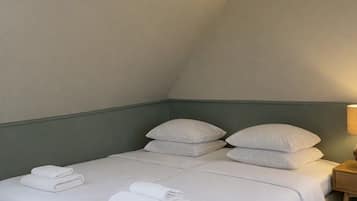 Una cama individual con sábanas y almohadas blancas, una mesita de noche de madera con una lámpara, y suelo cubierto de alfombra en una habitación con techo inclinado.