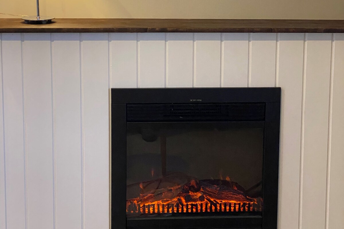Una chimenea con fuego a la vista, un cuadro colgado en la pared que muestra un puerto y una lámpara de mesa.