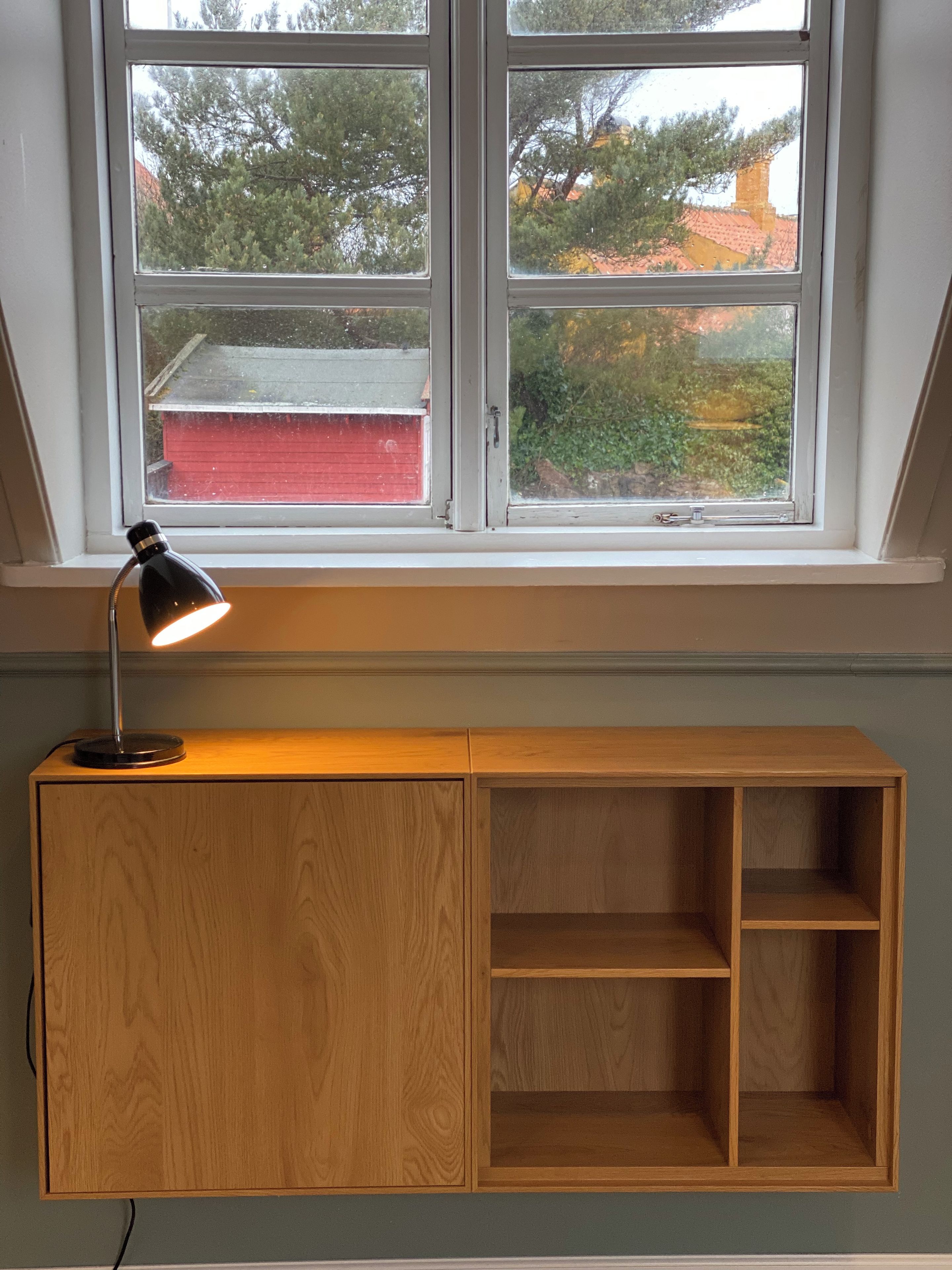 Una habitación con ventana, un escritorio con lámpara y vistas a árboles y edificios.