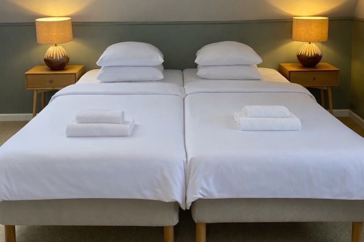 A room with two beds, each with a pillow and a rolled towel, two bedside tables with lamps, and a skylight above.