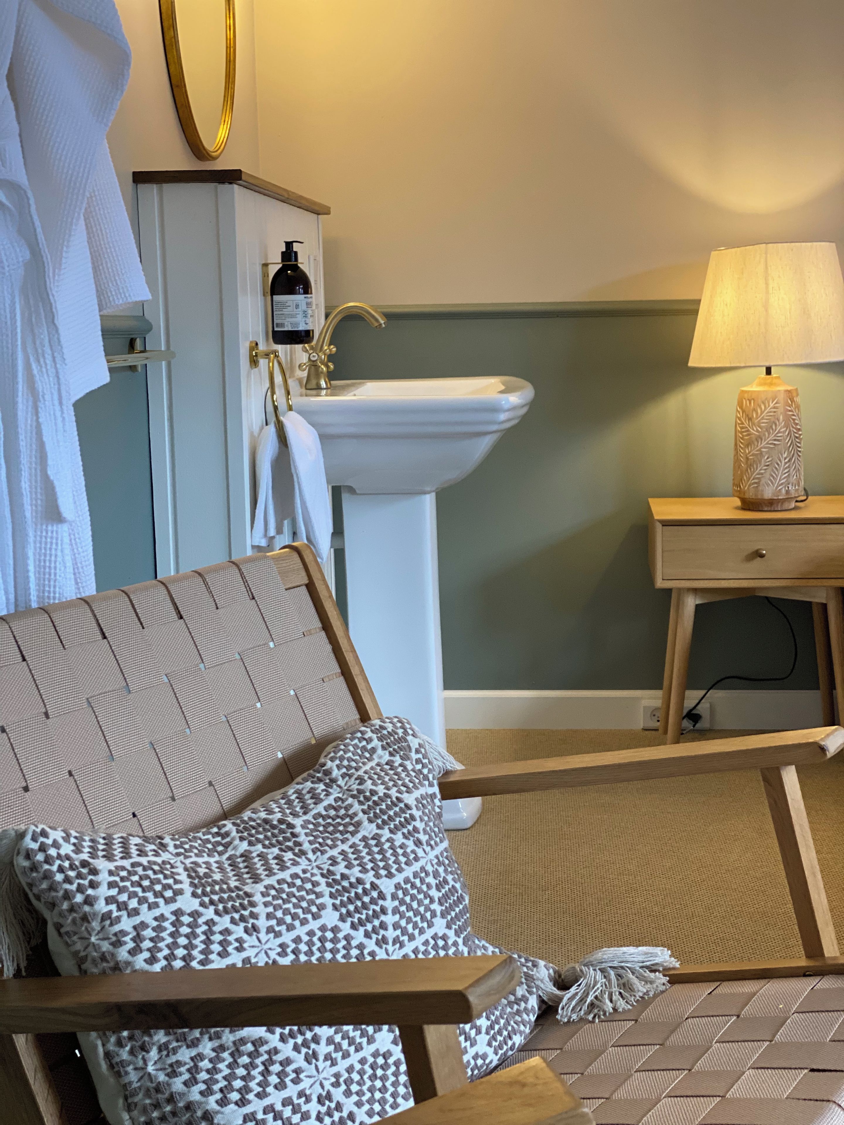 A hotel room with a bed, a bedside table with a lamp, a bathroom with a sink and mirror, and a towel rack.