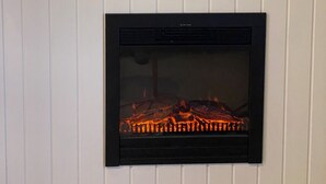 A fireplace with a visible fire, a wall-mounted picture of a harbor, and a table lamp. - Klippen - Skt. Jørgens Gaard (Gudhjem)