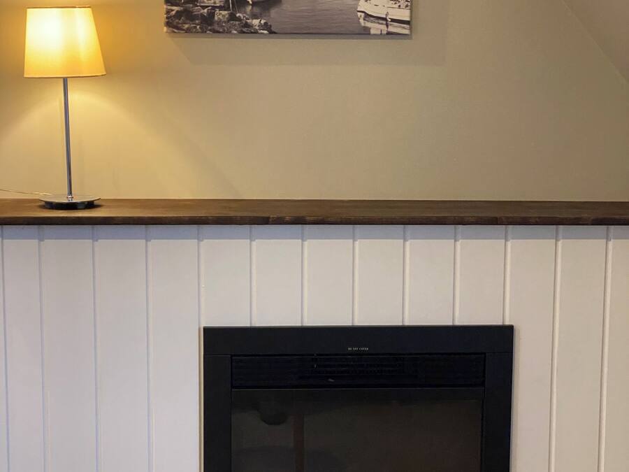 A fireplace with a visible fire, a wall-mounted picture of a harbor, and a table lamp.