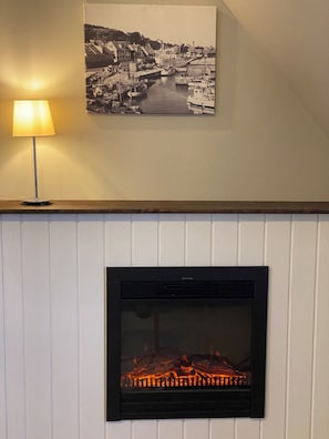 Une cheminée avec un feu apparent, un tableau représentant un port accroché au mur et une lampe de table.