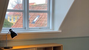 A room with a sloped ceiling, a window with a view of buildings, a desk with a lamp, and a chair. - Klippen - Skt. Jørgens Gaard (Gudhjem)