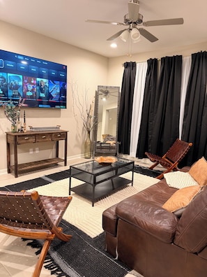 A living room with a flat-screen TV, a ceiling fan, a glass coffee table, a leather sofa, a wooden chair, and a mirror. - The House Mcallen 3bdroom Pool (McAllen)