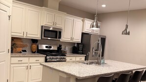A modern kitchen with white cabinetry, a granite countertop, stainless steel appliances, and bar stools. - The House Mcallen 3bdroom Pool (McAllen)