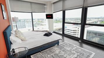 Una habitación de hotel moderna con una cama grande, una pantalla de televisión mostrando 'NETFLIX' y vistas de un paisaje urbano a través de amplios ventanales.