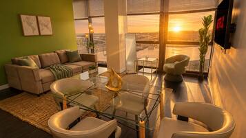 Una sala de estar moderna con un sofá, sillones, una mesa de centro de vidrio y vistas a un paisaje urbano a través de amplios ventanales.