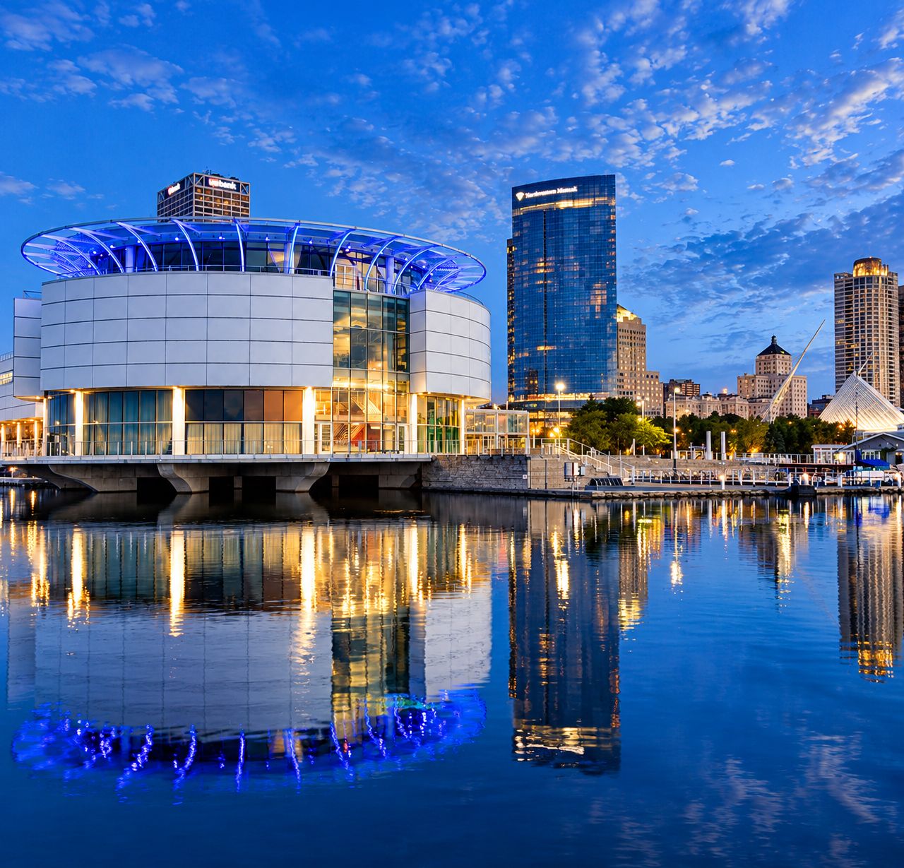 A waterfront building with a glass facade and a blue-lit structure, reflecting on the water.