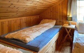 Ein Einzelbett in einem Holzraum mit schrägem Dach, ein Nachttisch mit Lampe und ein Fenster mit Vorhängen.