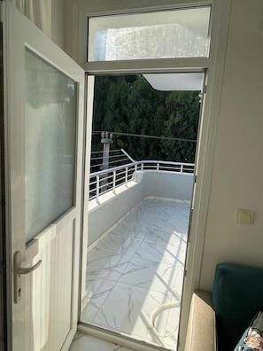 Una puerta abierta que da a un balcón con vistas a árboles, una barandilla y un suelo de baldosas.