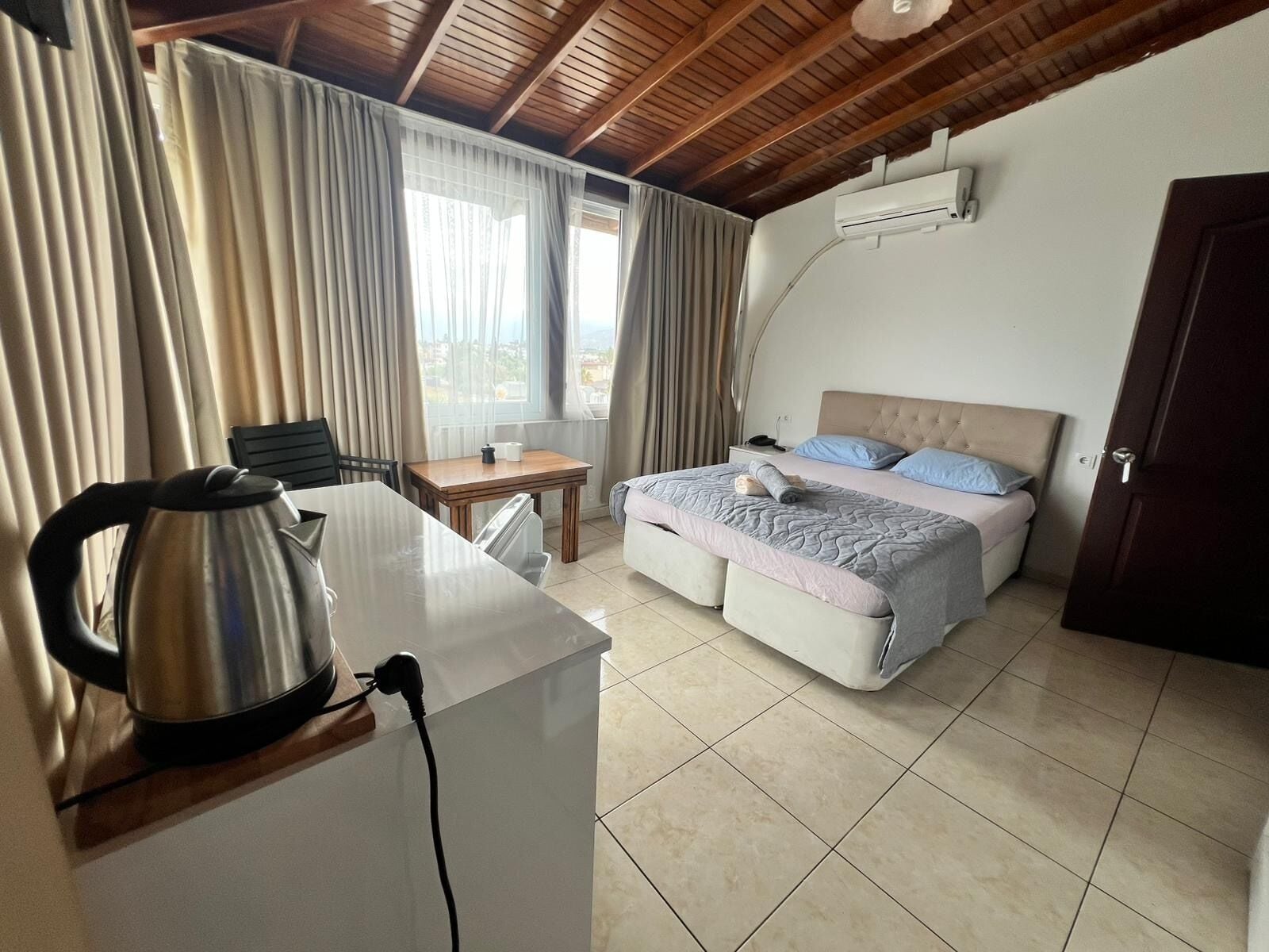 A hotel room with a bed, a desk with a kettle, a chair, a small table, and a window with curtains.