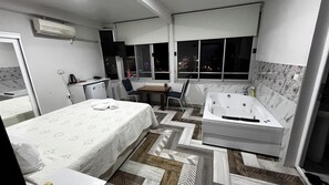 A hotel room with a bed, a small desk, a chair, a window with a view of the city at night, and a separate bathroom with a bathtub. - Arsuz Marin Otel (Arsuz)