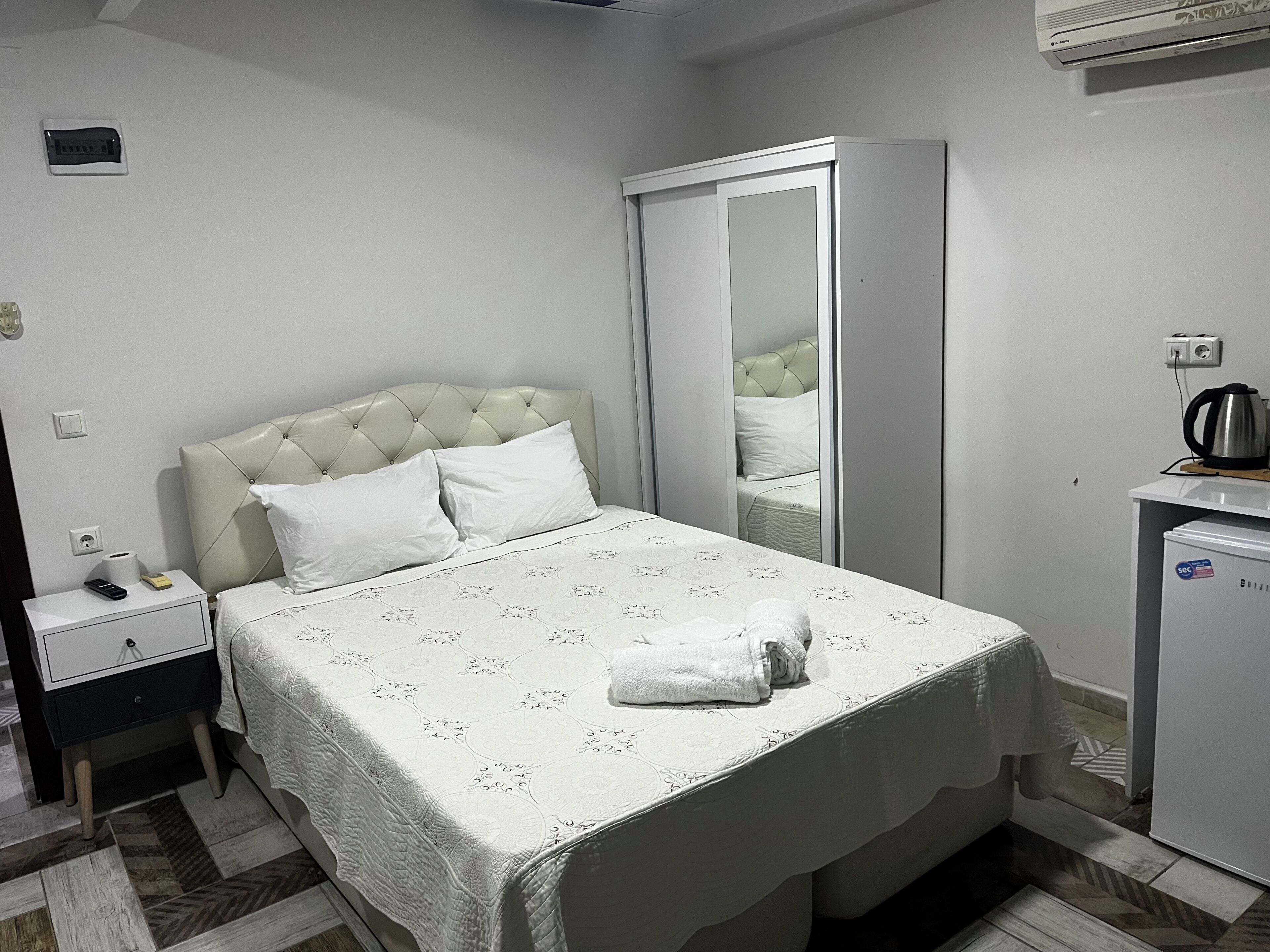 Una cama bien hecha con edredón blanco, una mesita de noche con control remoto, un armario pequeño y un espejo en la pared.