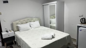 Una cama bien hecha con edredón blanco, una mesita de noche con control remoto, un armario pequeño y un espejo en la pared.