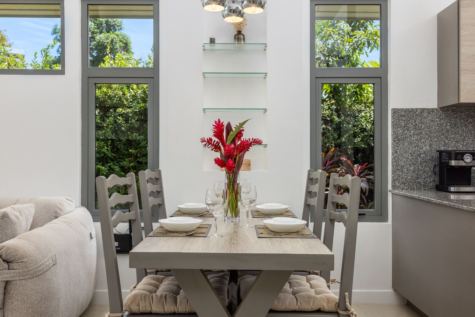 Un comedor con una mesa dispuesta para cuatro personas, rodeada de sillas y un florero con flores rojas sobre la mesa.
