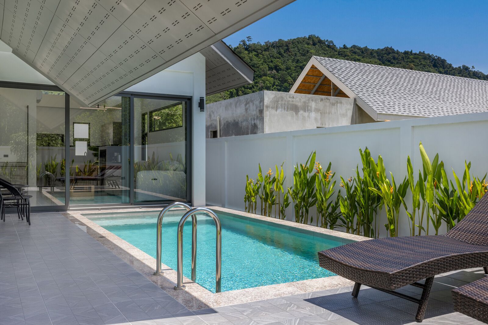 Un área de piscina moderna al aire libre con cerramiento de vidrio, sillones de descanso y vistas a una colina.