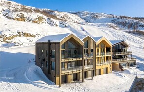 A modern wooden lodge with multiple balconies, set against a snowy mountainous backdrop.