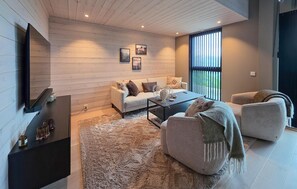 A modern living room with a sofa, coffee table, and armchairs, a flat-screen TV mounted on the wall, and a large window with blinds.