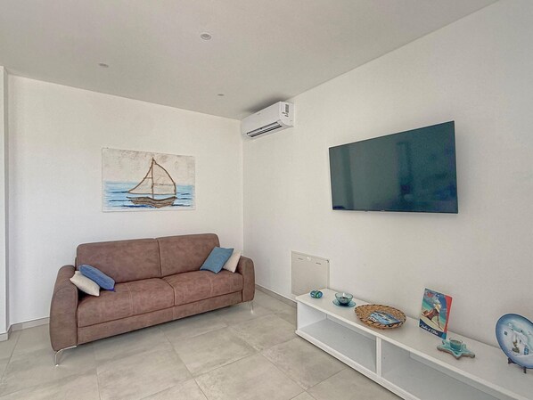 Ein Wohnzimmer mit einem braunen Sofa, einem Wand-Fernseher, einem Bild eines Segelbootes und einem Regal mit Dekorationsobjekten.