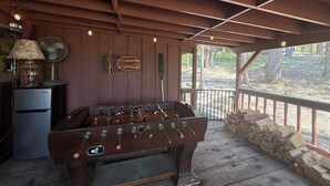 Une pièce au plancher de bois, équipée d’un baby-foot, d’un ventilateur ancien et d’un petit réfrigérateur.