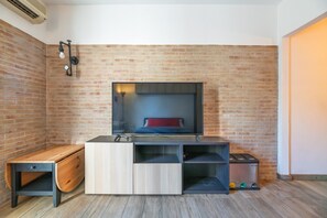 Una habitación con pared de ladrillo, un televisor sobre un soporte de madera, un banco y un cubo de basura.