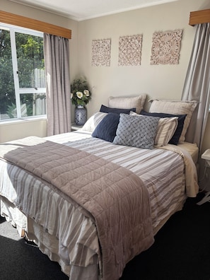 A bedroom with a large bed, a window with curtains, a vase with flowers, and wall art. - Lodge Ave Central Bach (Mount Maunganui)