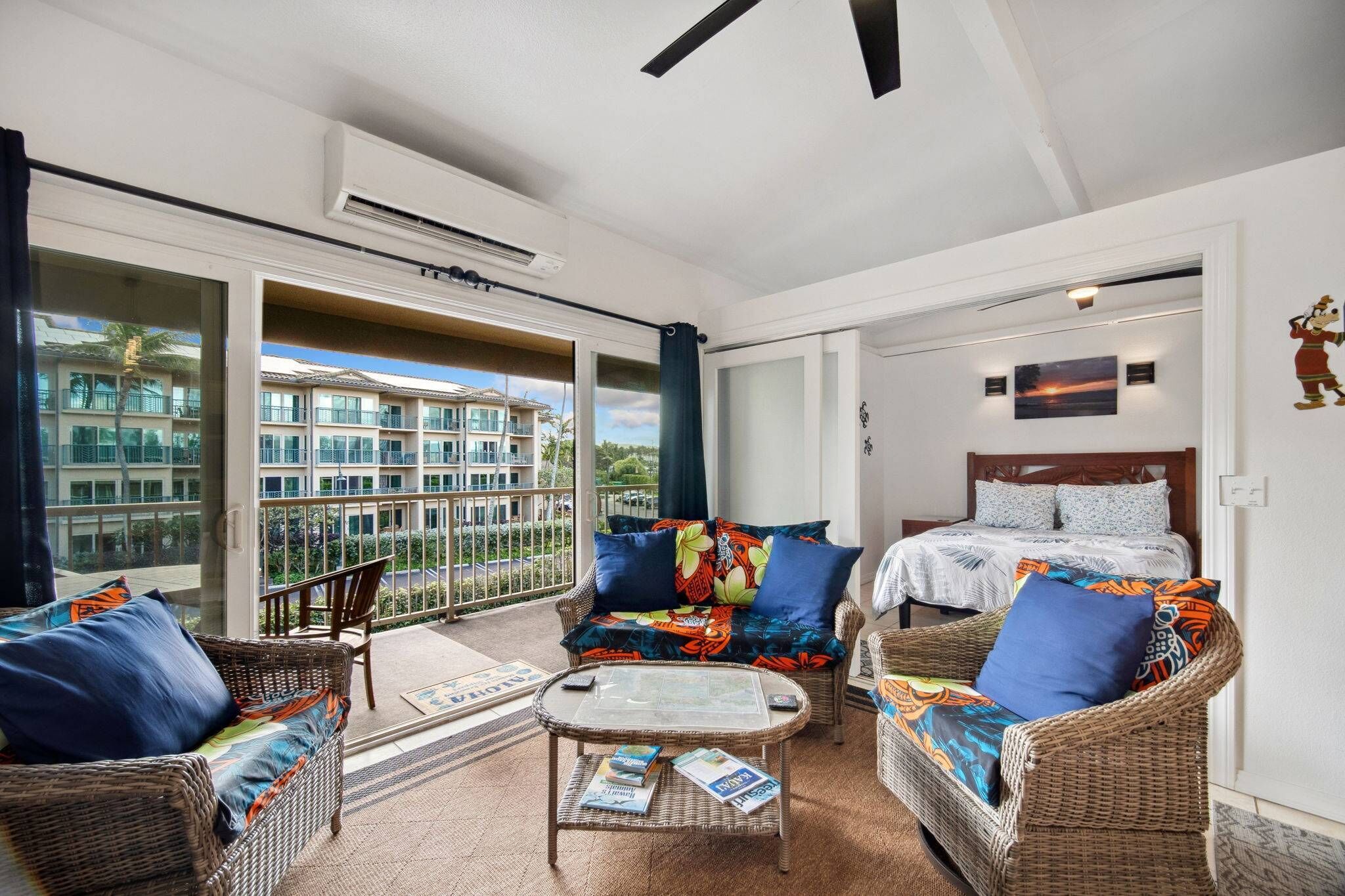 Varanda com vista para um edifício, móveis de vime, cama com roupa de cama estampada e um aparelho de ar condicionado.