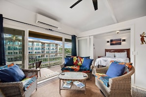 A balcony with a view of a building, wicker furniture, a bed with patterned bedding, and an air conditioning unit. - Cozy 2-bedroom condo centrally located in charming Kapaʻa with AC (Kapaʻa)