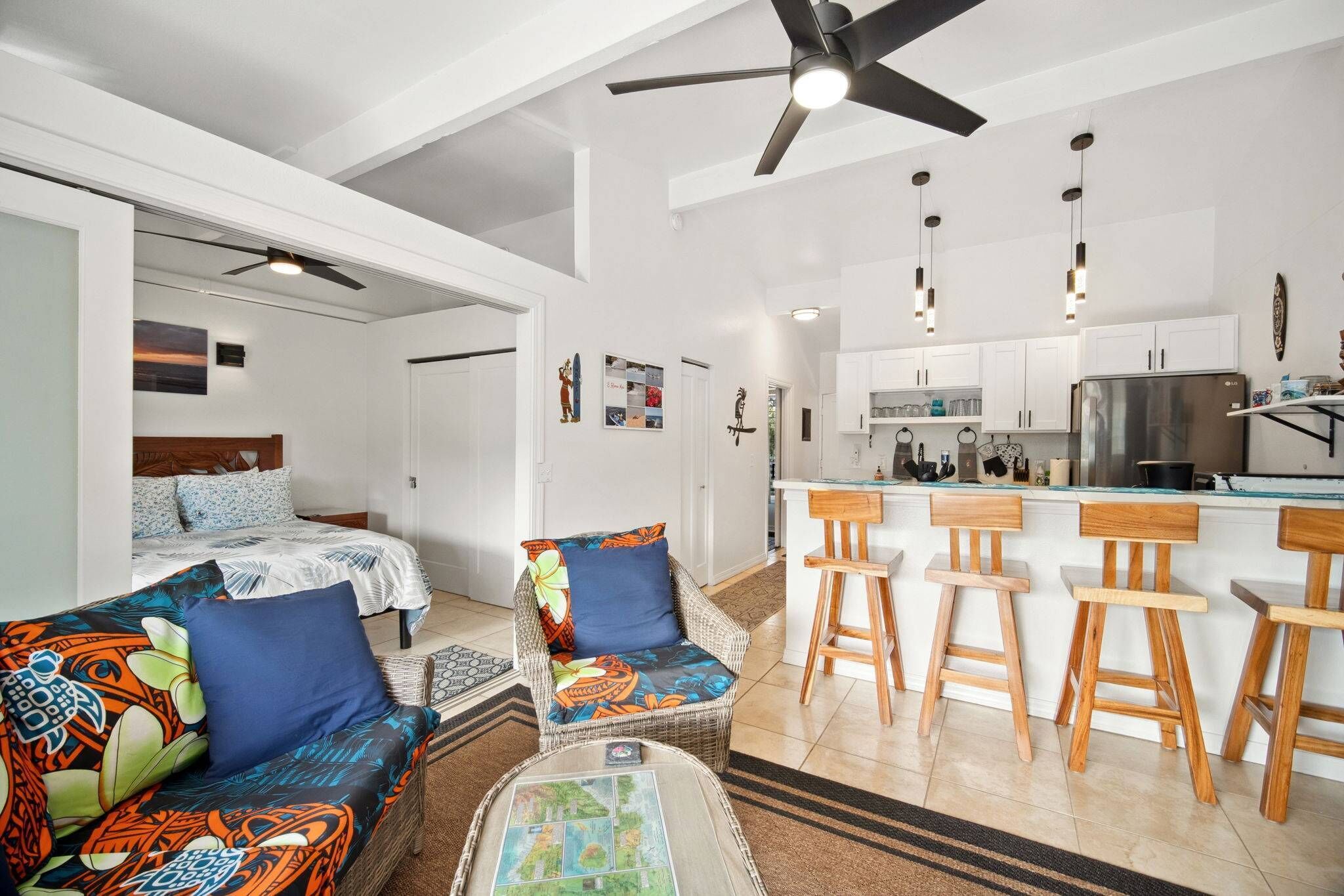 A modern living room with a sofa, a chair, a coffee table, a ceiling fan, and a kitchen area with a refrigerator and bar stools.