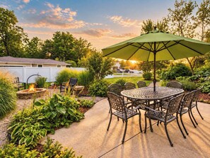Een terras buiten met een ronde tafel en stoelen, een vuurkorf en een groene parasol.