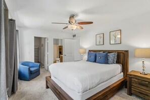 Une chambre à coucher comprenant un lit, une chaise, une table de chevet, une lampe, un ventilateur de plafond et une porte donnant sur une salle de bain.