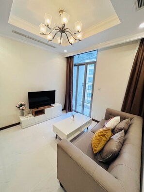 Ein modernes Wohnzimmer mit Flachbildfernseher, einem weißen Couchtisch, einem beige gefärbten Sofa mit Kissen und einer Deckenleuchte.