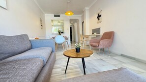A modern living room with a grey sofa, a round wooden coffee table, a pink armchair, and a dining area in the background.