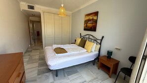 A bedroom with a bed, a nightstand, a chair, a painting, and a hallway leading to another room.