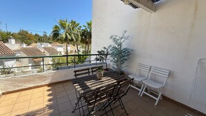 Ein Balkon mit Holztisch, zwei weißen Stühlen, einem Blumentopf und Blick auf Wohnhäuser.