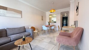 Ein modernes Wohnzimmer mit einem grauen Sofa, einem runden Holztisch, einem rosa Sessel und einem Essbereich mit Tisch und Stühlen.