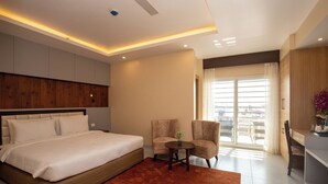 Ein modernes Hotelzimmer mit einem großen Bett, zwei Sesseln, einem kleinen Tisch und Blick ins Freie durch ein großes Fenster.