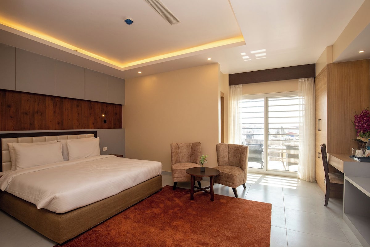 Una habitación de hotel moderna con una cama grande, dos sillones, una mesita y vistas al exterior a través de un ventanal.