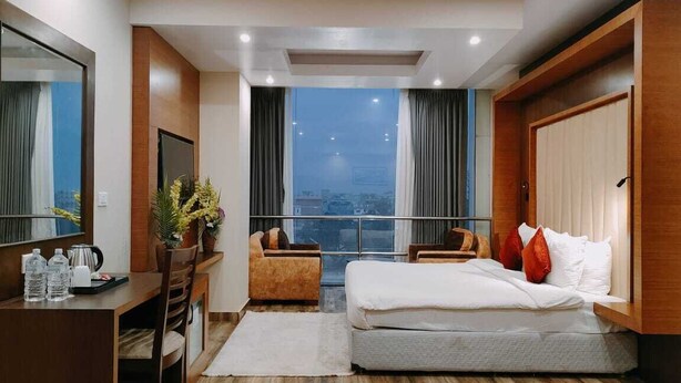 Ein Hotelzimmer mit Bett, Schreibtisch, Stuhl, einem großen Fenster mit Vorhängen und Blick auf die Stadt.