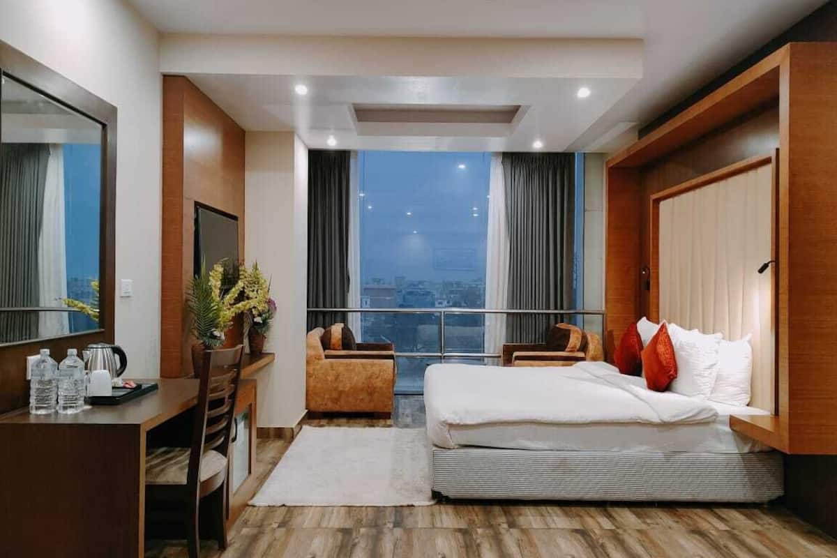 Una habitación de hotel con cama, escritorio, silla, una ventana grande con cortinas y vista a la ciudad.