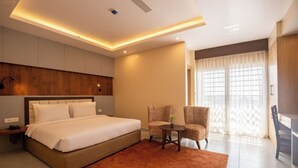 Ein modernes Hotelzimmer mit einem großen Bett, einer Sitzecke, einem kleinen Tisch und Blick ins Freie durch ein großes Fenster.