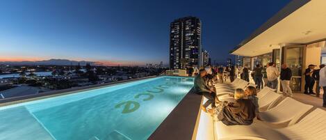 Un área de piscina en la azotea con vista a la ciudad, sillones de descanso y un grupo de personas.