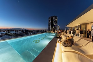 Un área de piscina en la azotea con vista a la ciudad, sillones de descanso y un grupo de personas.