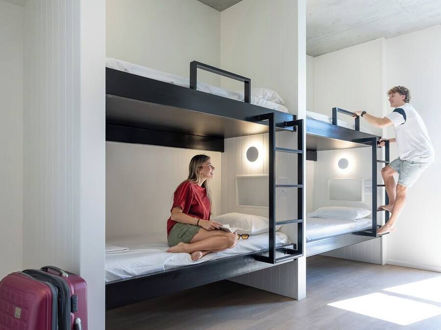 Habitación con literas, una maleta, una escalera y una persona escalando la escalera.
