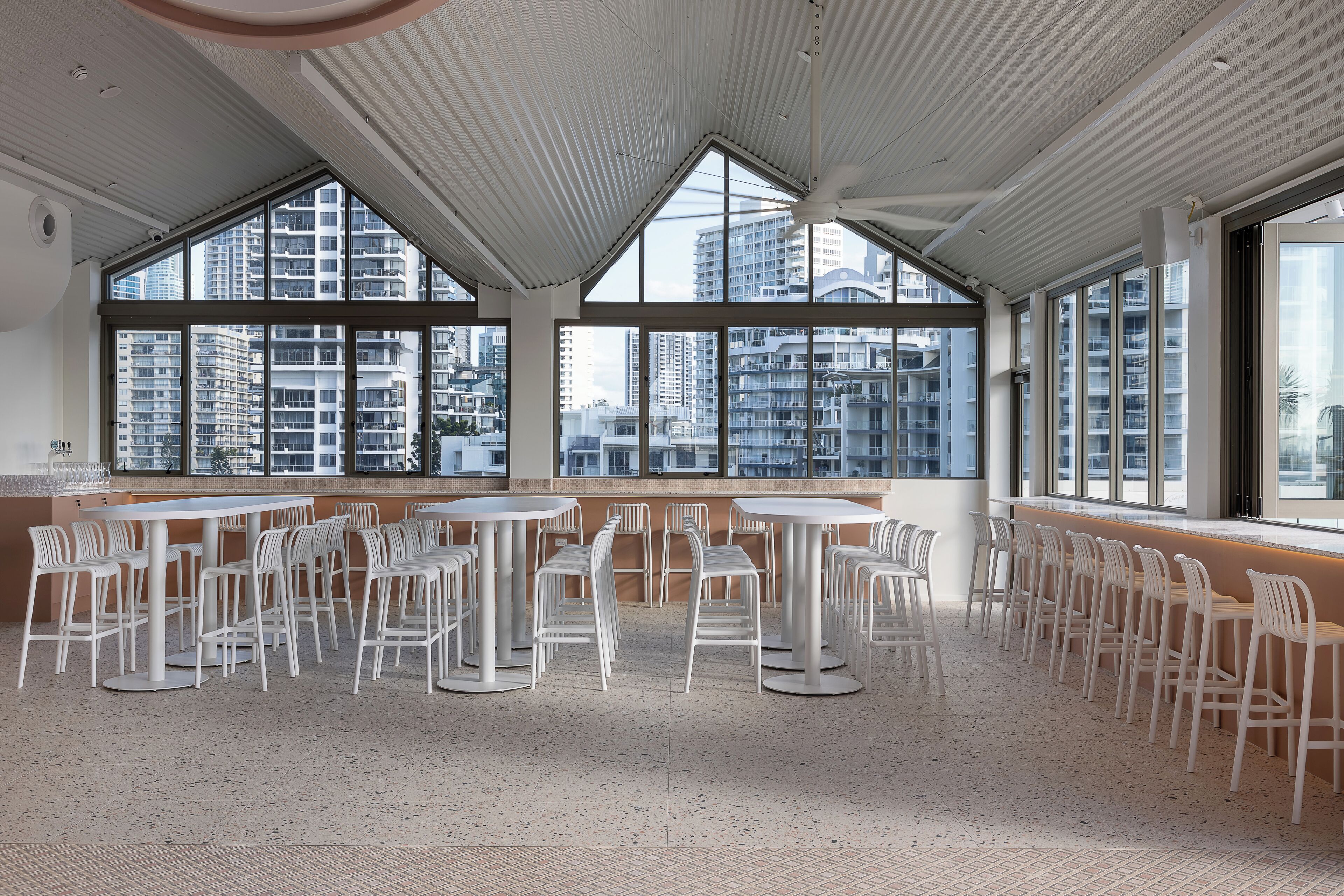 A spacious indoor area with multiple tables and chairs, large windows offering views of tall buildings, and a high ceiling with a ribbed design.