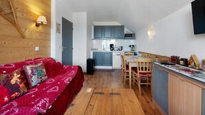 A cozy room with a wooden ceiling, a red sofa with festive pillows, a wooden dining table, and a kitchen area with cabinets and a microwave. - Apartment “Valmeinier Le Miras” with Mountain View, Private Terrace and Wi-Fi (Valmeinier)
