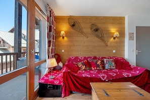 A cozy room with a wooden wall, two wall-mounted lamps, a red-covered sofa with festive pillows, and a wooden bench. - Apartment “Valmeinier Le Miras” with Mountain View, Private Terrace and Wi-Fi (Valmeinier)